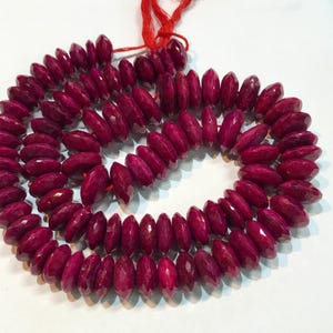 May include: A strand of deep red ruby beads, each bead is faceted and has a slightly oval shape. The beads are strung on a red thread.