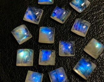 Könnte beinhalten: Eine Sammlung von 14 regenbogenfarbenen Mondstein-Cabochons, die jeweils in quadratischer Form geschnitten sind. Die Steine sind milchig weiß mit einem blauen Schimmer.