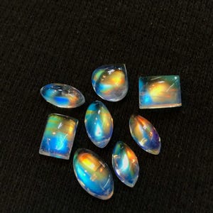 Puede incluir: Una colecci&oacute;n de siete cabujones de piedra de luna arco&iacute;ris iridiscentes en varias formas y tama&ntilde;os. Las piedras est&aacute;n colocadas sobre un fondo negro.