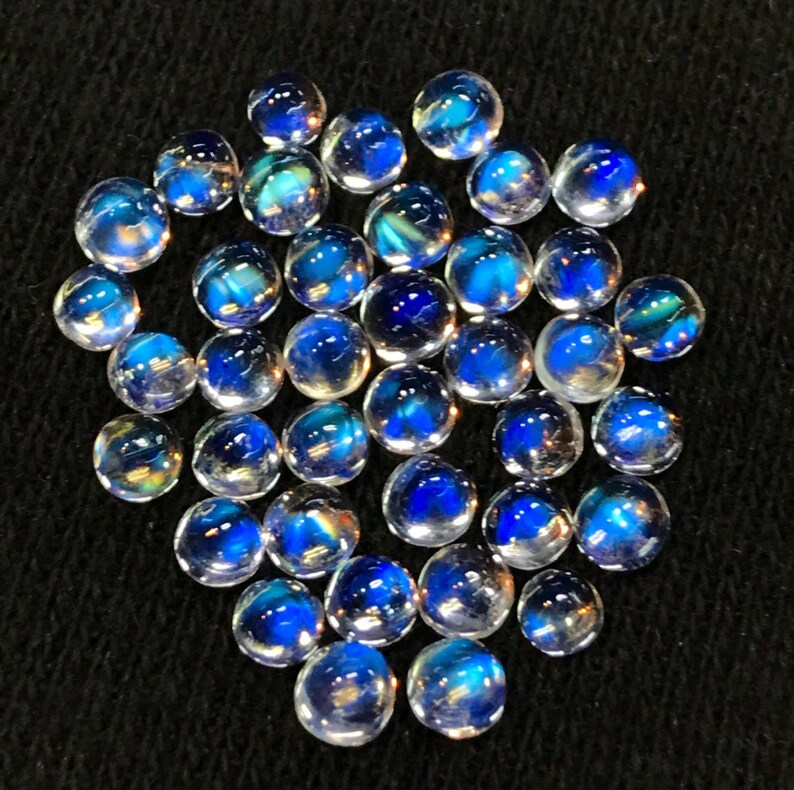 Op de afbeelding: Een verzameling van iriserende blauwe en witte edelsteen cabochons. De cabochons zijn rond en hebben een glad, gepolijst oppervlak. Ze zijn in een losse stapel op een zwarte achtergrond gerangschikt.