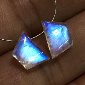 May include: Two iridescent, kite-shaped moonstone gemstones. The stones display a vibrant blue and white colour with internal light reflections. They are strung on a thin, silver-coloured wire, ready for jewellery making.