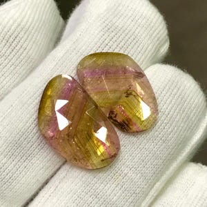 May include: Two faceted gemstones with a mix of yellow, pink, and gold hues. The stones have an oval shape and are set against a white fabric background. The gems have a polished, reflective surface.