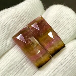 May include: Two rectangular gemstones with faceted edges. The stones display a gradient of colors, including pink, yellow, and brown. The gems are set against a white fabric background, highlighting their polished surfaces and intricate cuts.