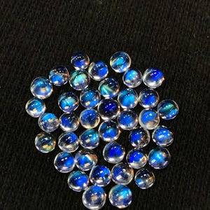 Op de afbeelding: Een verzameling ronde, iriserende edelsteencabochons in verschillende tinten blauw. De cabochons zijn glad en gepolijst, met een doorschijnende uitstraling. Ze zijn in een losse stapel op een zwarte achtergrond gerangschikt.