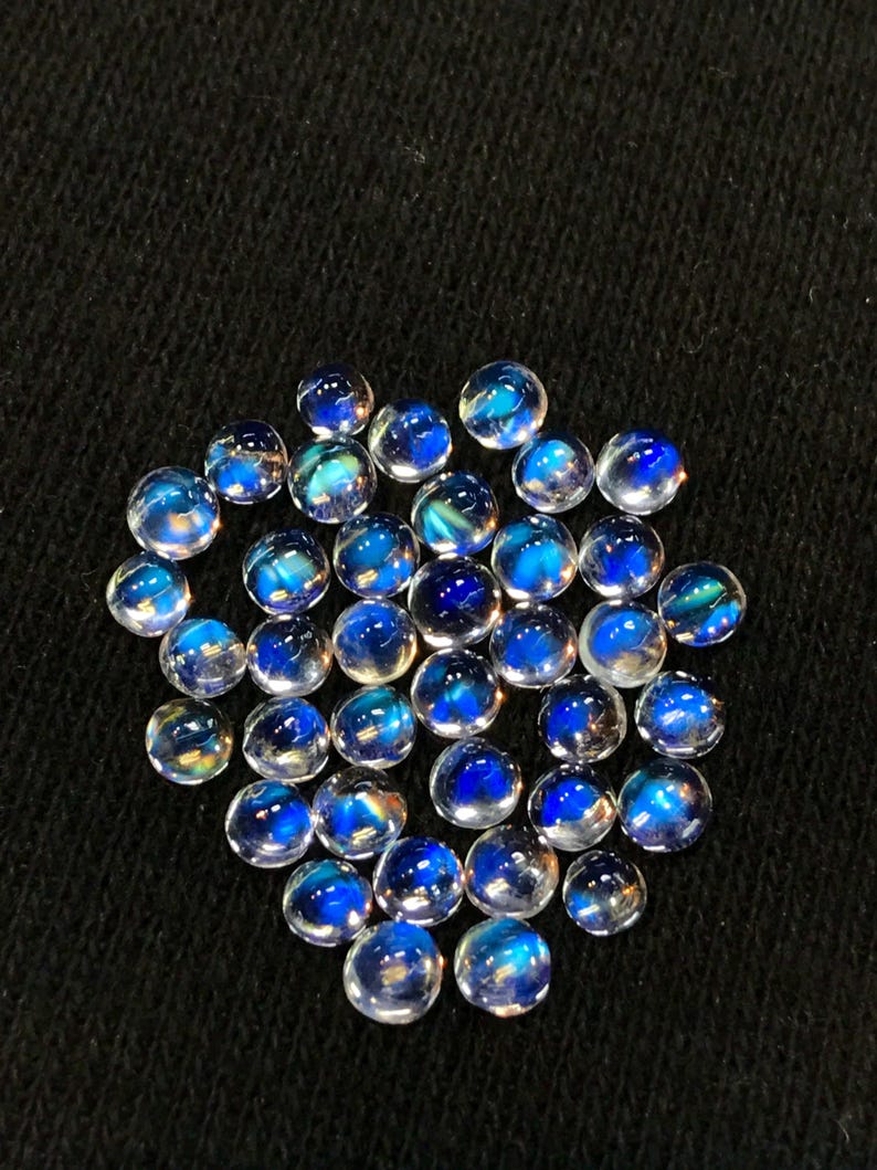 Op de afbeelding: Een verzameling ronde, iriserende edelsteencabochons in verschillende tinten blauw. De cabochons zijn glad en gepolijst, met een doorschijnende uitstraling. Ze zijn in een losse stapel op een zwarte achtergrond gerangschikt.