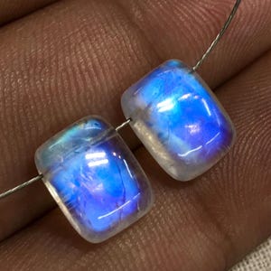 May include: Two iridescent blue and white moonstone beads strung on a silver wire. The beads are square shaped and have a smooth, polished finish.