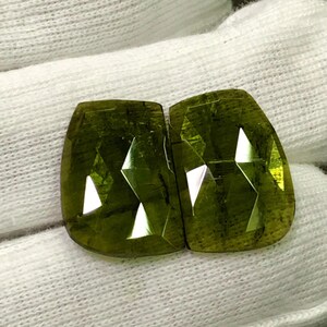 May include: Two faceted, olive-green gemstones with a rectangular shape and rounded corners. The gems have multiple facets that reflect light, creating bright white triangular shapes. The stones are displayed on a white, textured surface.