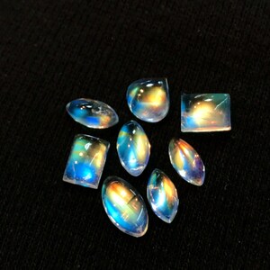 Puede incluir: Una colecci&oacute;n de ocho cabujones de piedras preciosas iridiscentes en varias formas y tama&ntilde;os. Las piedras tienen un brillo arco&iacute;ris y se muestran sobre un fondo negro.