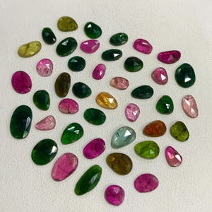 May include: A collection of green, pink, and brown gemstones in various shapes and sizes. The gemstones are arranged in a heart shape on a white surface.