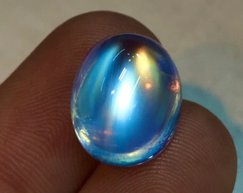 May include: An oval-shaped, iridescent gemstone with a blue and yellow color gradient. The polished surface reflects light, creating a shimmering effect. The gemstone is smooth and appears to be a cabochon cut.