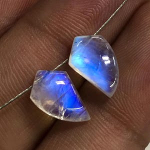 May include: Two polished, iridescent moonstone gemstones with a blue and white colour scheme. The gemstones are cut into a unique shape and strung on a thin, silver-coloured wire. The stones have a translucent appearance.
