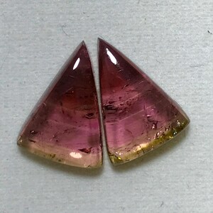 May include: Two triangular shaped gemstones with a pink, green, and yellow color gradient. The gemstones are cut into a sharp point at the top and have a smooth, polished surface.