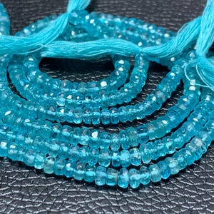 Op de afbeelding: Een streng gefacetteerde, doorschijnende blauwe apatietkralen gerangschikt in een cirkelvormig patroon. De kleine, ronde, schijfvormige kralen hebben een levendige cyaan kleur en zijn geregen aan een helder turquoise draad met een kwastje.
