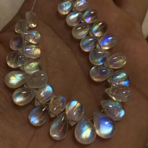 May include: Strands of iridescent moonstone beads. The teardrop-shaped stones display a play of colour, with flashes of blue, yellow, and green. The beads are strung together, ready for jewellery making. The stones are translucent and have a polished finish.