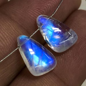 May include: Two polished, teardrop-shaped moonstone beads. The beads are translucent with a milky white base and vibrant blue and purple internal reflections. Strung on a thin, silver-coloured wire, they are ready for jewellery making.