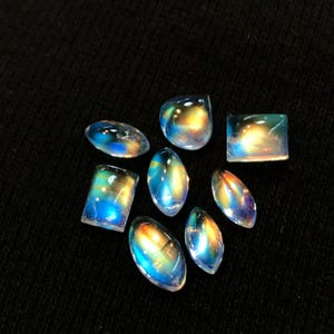 Puede incluir: Una colecci&oacute;n de ocho cabujones de piedras preciosas iridiscentes en varias formas y tama&ntilde;os. Las piedras son una mezcla de formas ovaladas, de l&aacute;grima y rectangulares. Los cabujones son de un blanco transl&uacute;cido con un brillo arco&iacute;ris.