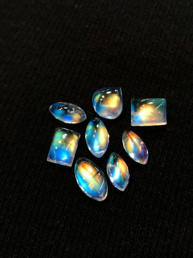 Puede incluir: Una colecci&oacute;n de ocho cabujones de piedras preciosas iridiscentes en varias formas y tama&ntilde;os. Las piedras son una mezcla de formas ovaladas, de l&aacute;grima y rectangulares. Los cabujones son de un blanco transl&uacute;cido con un brillo arco&iacute;ris.