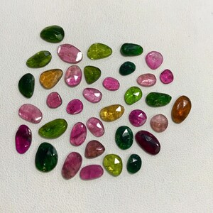 May include: A collection of 25 faceted gemstones in various shades of green, pink, and orange. The gemstones are cut in a freeform shape and have a smooth, polished finish.