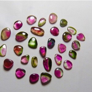 May include: A collection of 25 loose, natural, multi-colored gemstones. The stones are a mix of pink, green, and black, and are all different shapes and sizes.