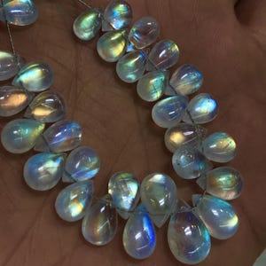 May include: A collection of iridescent moonstone teardrop beads. The beads are strung on a thin wire, displaying a spectrum of colours including blue, green, and yellow. The beads are smooth and polished, reflecting light.