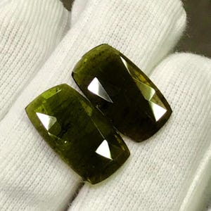May include: Two faceted, olive-green gemstones with rectangular shapes. The gems have multiple facets that reflect light, creating bright, triangular highlights. The stones are displayed on a white, textured surface.