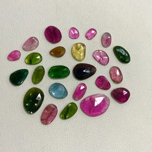 May include: Assortment of 20 loose gemstones in various colors, including pink, green, blue, and yellow. The gemstones are cut in a variety of shapes, including oval, round, and pear.