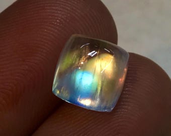 May include: A rainbow moonstone cabochon gemstone with a light blue, green, and yellow iridescent sheen.