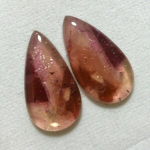 May include: Two teardrop-shaped gemstones with pink, red, and orange hues. The gemstones are translucent and have a sparkly, iridescent finish.