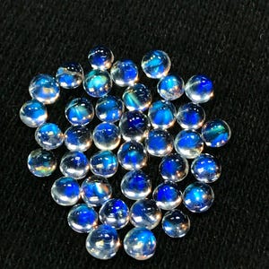 Op de afbeelding: Een verzameling ronde, iriserende maansteen cabochons met een blauwe glans. De stenen zijn ongeveer 6 mm in diameter en zijn in een losse stapel gerangschikt.