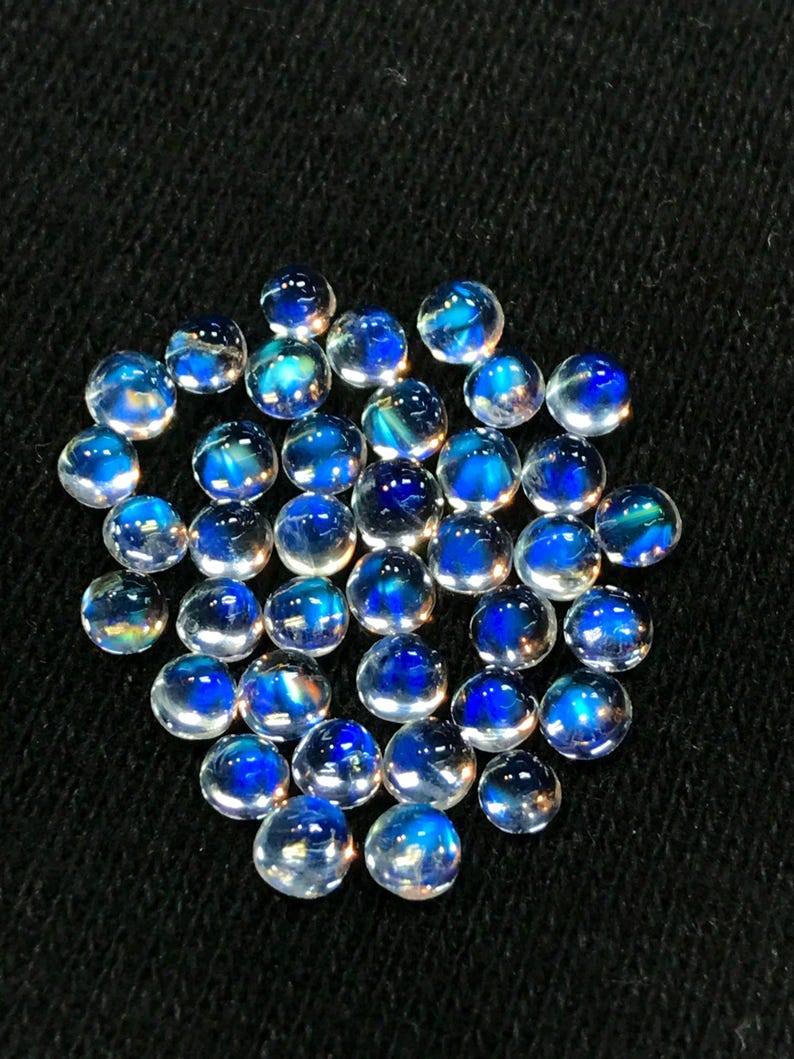 Op de afbeelding: Een verzameling ronde, iriserende maansteen cabochons met een blauwe glans. De stenen zijn ongeveer 6 mm in diameter en zijn in een losse stapel gerangschikt.