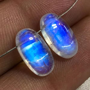 May include: Two oval-shaped moonstone beads with an iridescent blue and white colour.