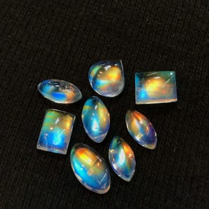 Puede incluir: Una colecci&oacute;n de ocho cabujones de piedras preciosas iridiscentes en varias formas y tama&ntilde;os. Las piedras son una mezcla de formas ovaladas, redondas y rectangulares. Los cabujones son de color azul claro con un brillo arco&iacute;ris.