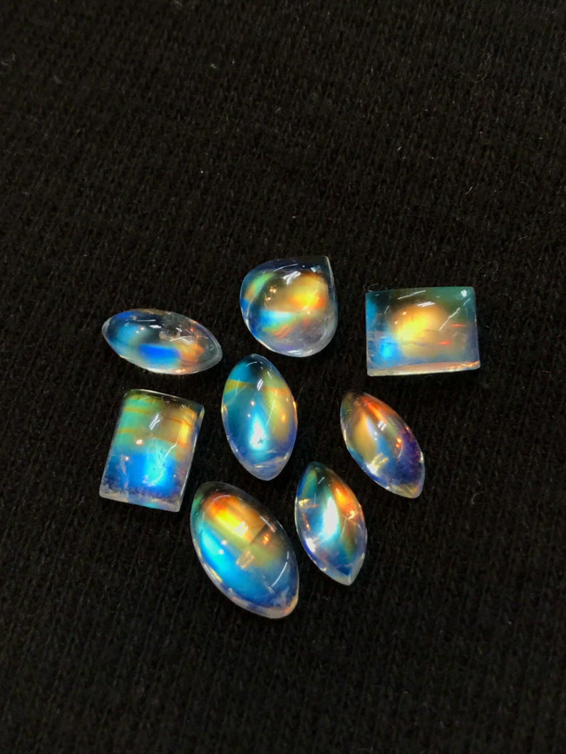 Puede incluir: Una colecci&oacute;n de ocho cabujones de piedras preciosas iridiscentes en varias formas y tama&ntilde;os. Las piedras son una mezcla de formas ovaladas, redondas y rectangulares. Los cabujones son de color azul claro con un brillo arco&iacute;ris.