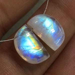 May include: Two polished moonstone cabochons, each with a semi-circular shape. The stones display a milky white base with iridescent flashes of blue, yellow, and purple. They are strung on a thin, silver-coloured wire, ready for jewellery making.