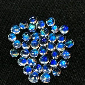 Op de afbeelding: Een verzameling ronde, iriserende maansteen cabochons met een blauwe schijn. De stenen zijn ongeveer 6 mm in diameter en zijn in een losse stapel gerangschikt.