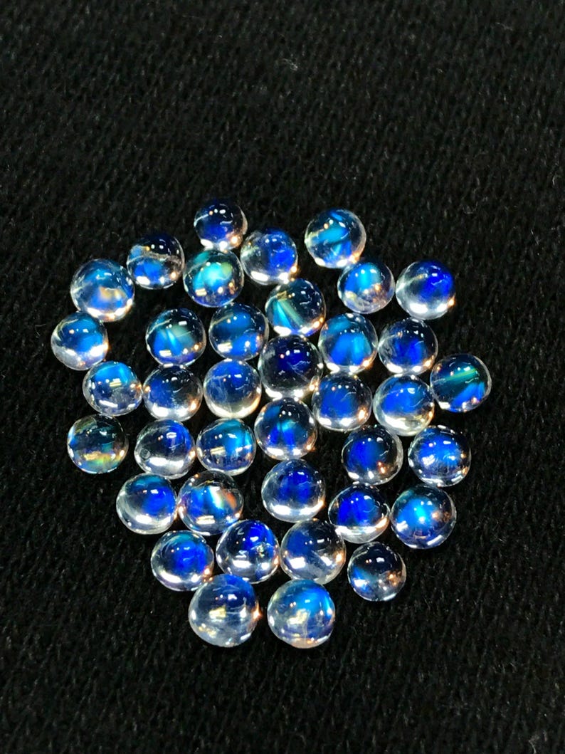 Op de afbeelding: Een verzameling ronde, iriserende maansteen cabochons met een blauwe schijn. De stenen zijn ongeveer 6 mm in diameter en zijn in een losse stapel gerangschikt.