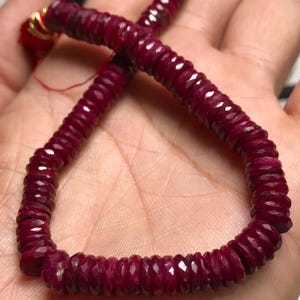 May include: A strand of deep red ruby beads, each bead is faceted and has a slightly elongated shape. The beads are strung on a thin cord and are displayed on a hand.