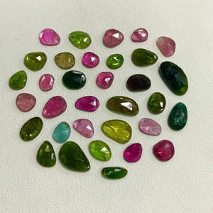 May include: A collection of faceted gemstones in various shapes and sizes. The gems display a range of colors, including pink, green, yellow, and teal. The stones are arranged on a white surface, showcasing their polished surfaces and internal clarity.