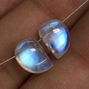 May include: Two translucent moonstone beads, polished to a smooth finish, with a blue and white iridescent sheen. They are strung on a thin, silver-coloured wire, highlighting their rounded shape. These gemstones are suitable for jewellery making.