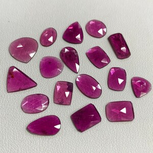 May include: Assortment of 15 pink gemstones, cut in various shapes, including oval, pear, and square. The gemstones are faceted and have a translucent appearance.