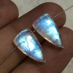 May include: Two polished, triangular moonstone beads with a translucent, iridescent quality. The beads are strung on a thin, silver-coloured wire, showcasing their natural blue and white hues. These gemstones are ideal for jewellery making.
