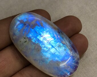Könnte beinhalten: Ein großer, ovaler Mondstein-Cabochon mit einem blauen, irisierenden Schimmer. Der Stein ist durchscheinend und hat eine weiße Grundfarbe.