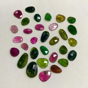 May include: Assortment of green, pink, and brown gemstones in various shapes and sizes. The gemstones are arranged in a heart shape on a white surface.