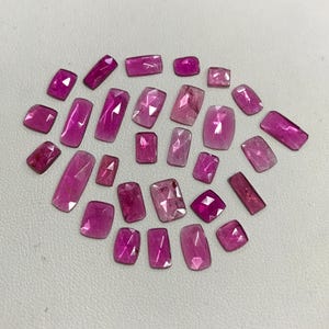 May include: A collection of 20 pink and purple gemstones, cut into rectangular shapes. The gemstones are arranged in a loose cluster on a white surface.