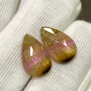 May include: Two faceted, teardrop-shaped gemstones. The gems have a color gradient, transitioning from a golden-brown hue to a pinkish-purple. The gems are displayed on a white, textured surface.