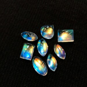 Puede incluir: Una colecci&oacute;n de ocho cabochones de piedra de luna arco&iacute;ris iridiscentes en varias formas y tama&ntilde;os. Las piedras est&aacute;n colocadas sobre un fondo de tela negra.