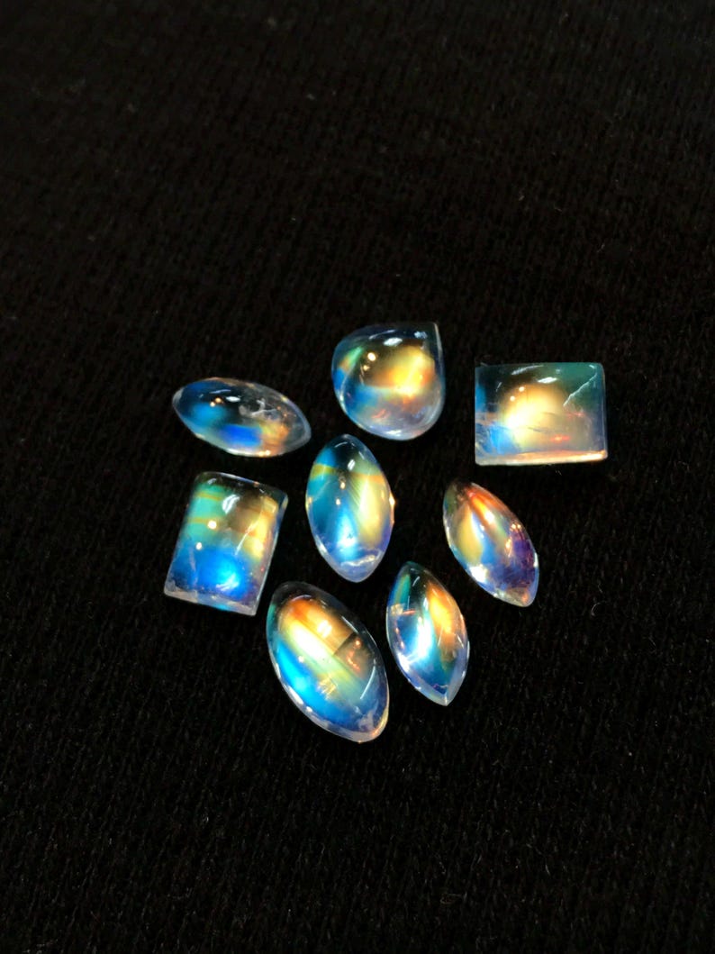 Puede incluir: Una colecci&oacute;n de ocho cabochones de piedra de luna arco&iacute;ris iridiscentes en varias formas y tama&ntilde;os. Las piedras est&aacute;n colocadas sobre un fondo de tela negra.