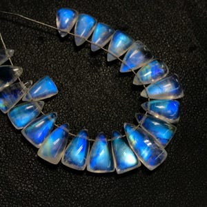 May include: A strand of iridescent blue moonstone beads, each bead is shaped like a teardrop and has a pointed tip. The beads are strung on a thin wire and are arranged in a circle.