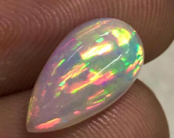 Könnte beinhalten: Ein birnenförmiger Opal-Edelstein mit milchig-weißer Basis und lebendigen Farbblitzen. Der Opal zeigt ein Farbspiel mit Farbtönen von Gelb, Grün, Rosa und Orange. Der Edelstein wird zwischen zwei Fingern gehalten.
