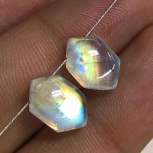 May include: Two faceted, iridescent moonstone beads strung on a thin, silver-coloured wire. The beads have a translucent quality with flashes of blue, yellow, and green. The gemstones are hexagonal in shape.